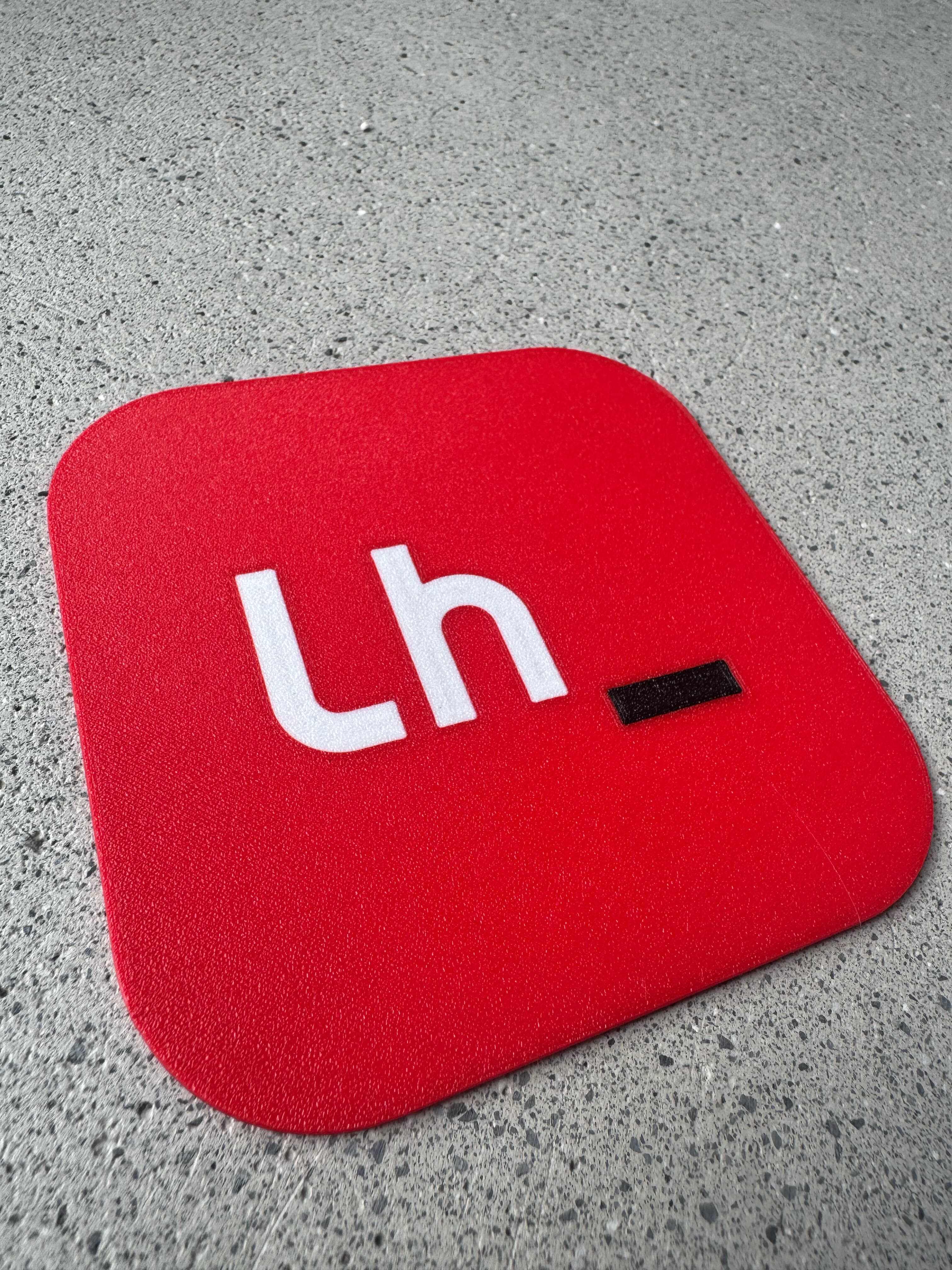 Red square coaster with a white “Lh” and a black cursor, 3D printed in three colors, on a terrazzo floor